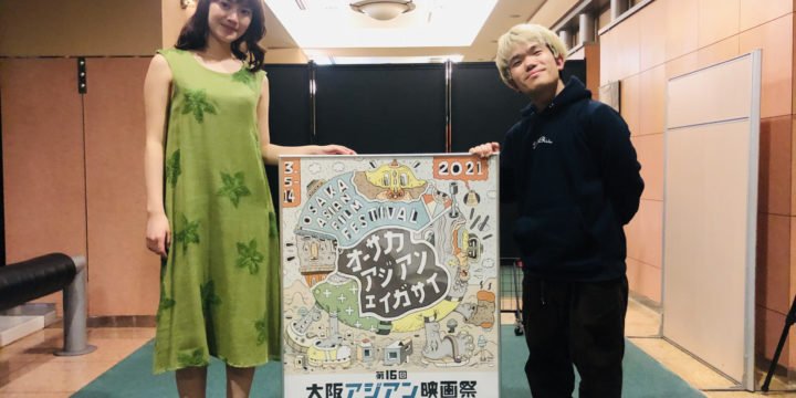 DirAct workshop ディラクト ワークショップ - 緑のワンピースの女性と黒いパーカーの男性が映画祭ポスターを前に笑顔の記念写真、スクールと演技の雰囲気を伝える場面
